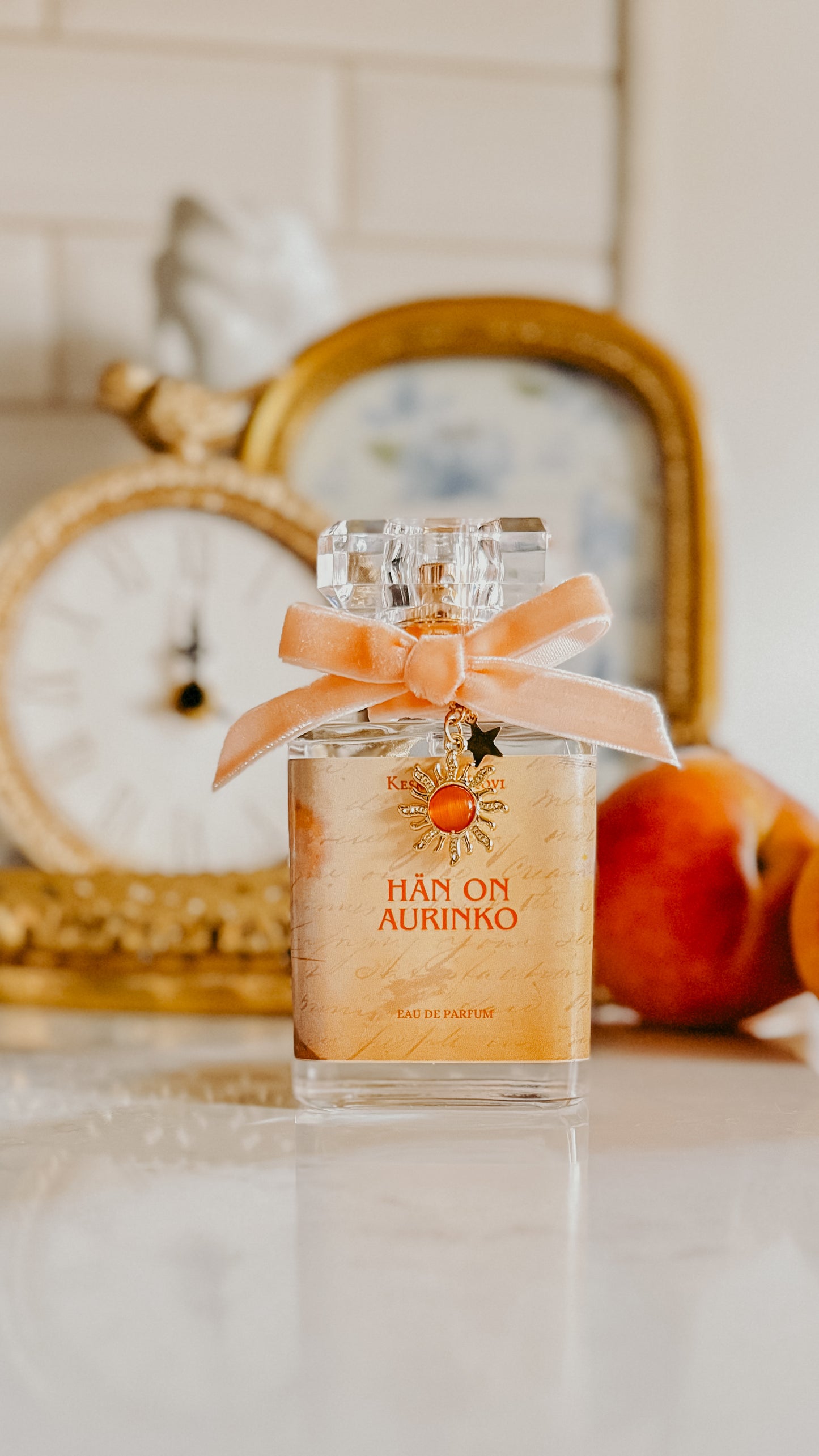 Perfume bottle with an orange ribbon on a surface with a decorative mirror and fruit in the background.
