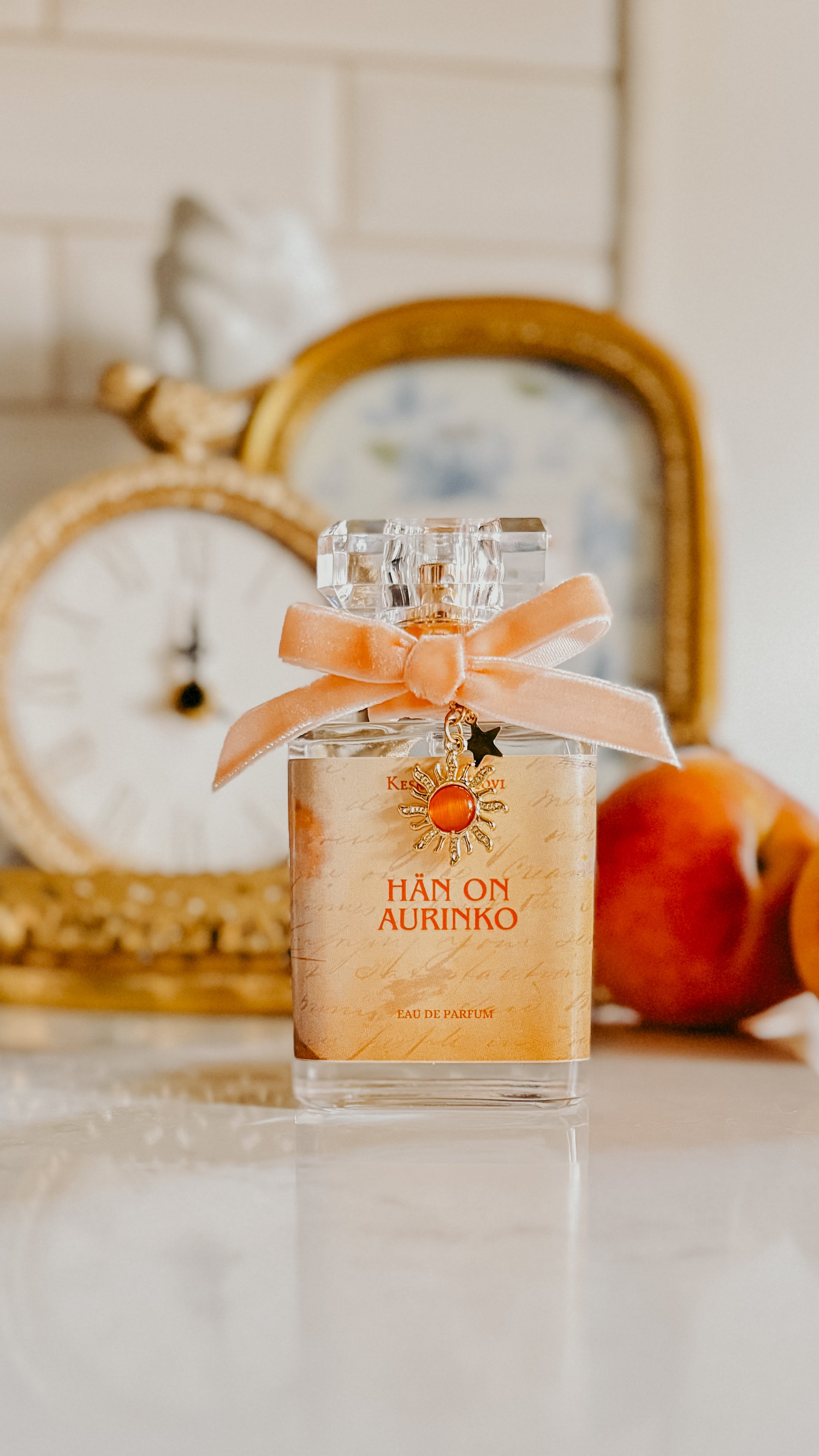 Perfume bottle with an orange ribbon on a surface with a decorative mirror and fruit in the background.