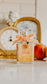 Perfume bottle with an orange ribbon on a surface with a decorative mirror and fruit in the background.