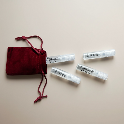 Four small perfume bottles with labels next to a red velvet bag on a beige background