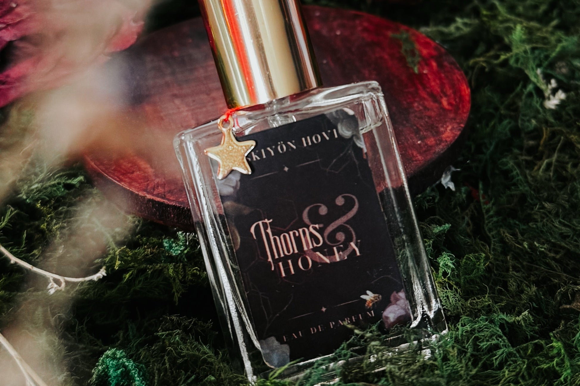 A clear rectangular perfume bottle with a shiny gold cap and a black label reading 'Thorns & Honey' by Kydon Home, styled on green moss with dried red flowers and a blurred natural background.